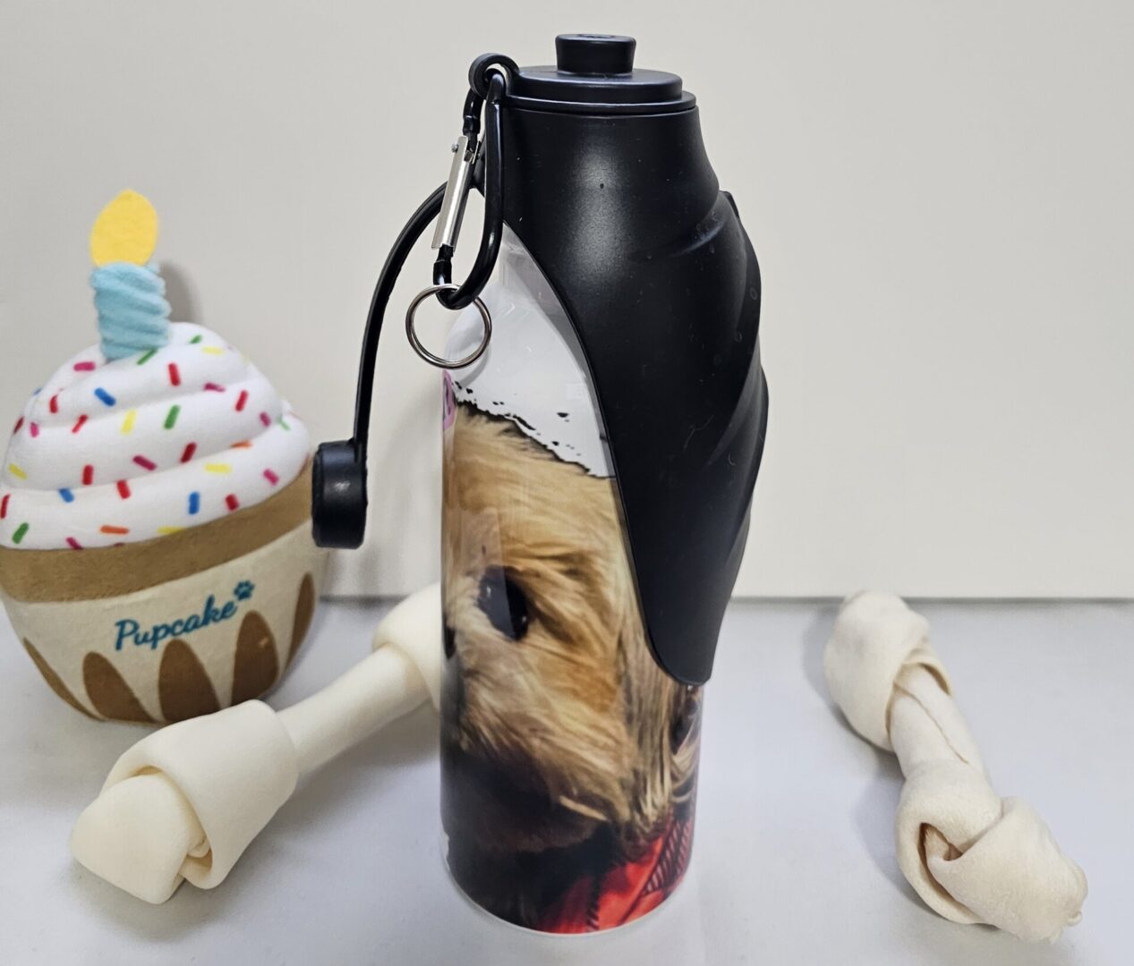 A 20 oz. Pet Water Bottle with an image of a dog on it, flanked by a plush cupcake toy and two rawhide bones.