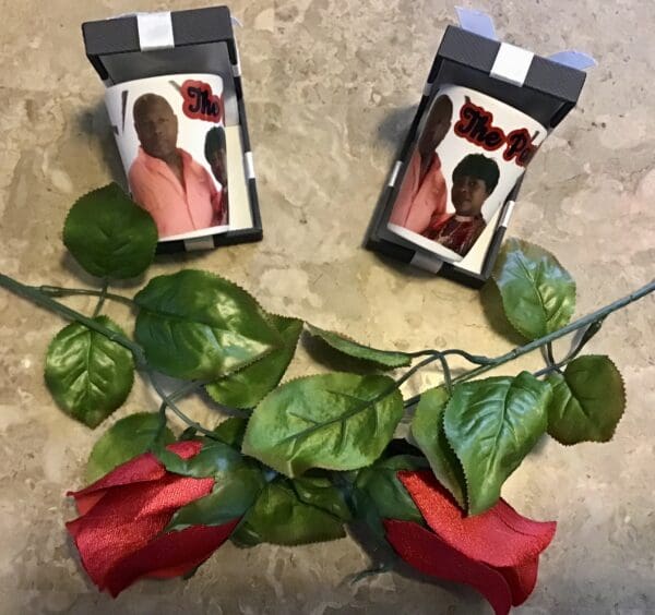 Two 1.5 oz. Ceramic Shot Glasses with a photo of a man on them, lying next to a stem of artificial red roses.
