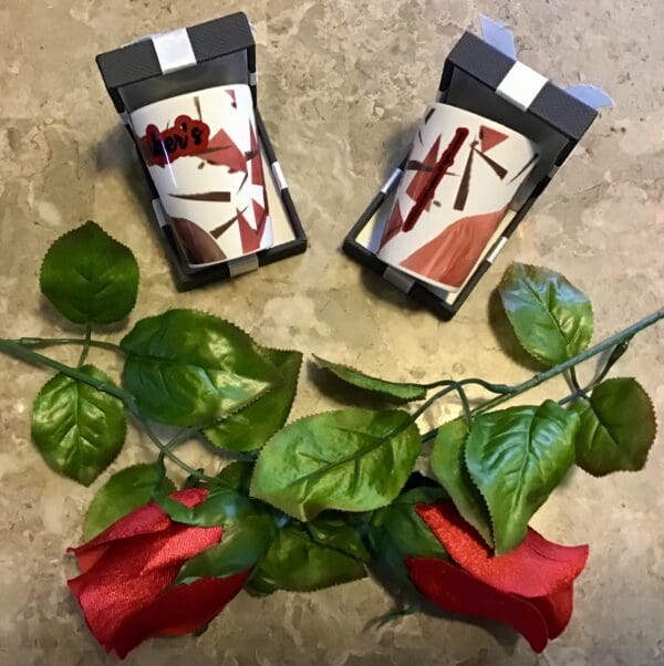 Two 1.5 oz. Ceramic Shot Glasses with heart patterns and artificial roses lying on a textured surface.